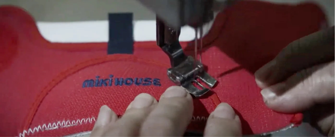 A craftsperson stitches a red MIKI HOUSE shoe upper